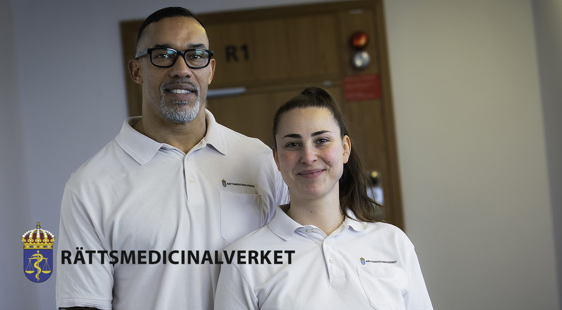 Två medarbetare står bredvid varandra inomhus och bär vita pikétröjor med myndighetens logotyp. Rättsmedicinalverkets logotyp.