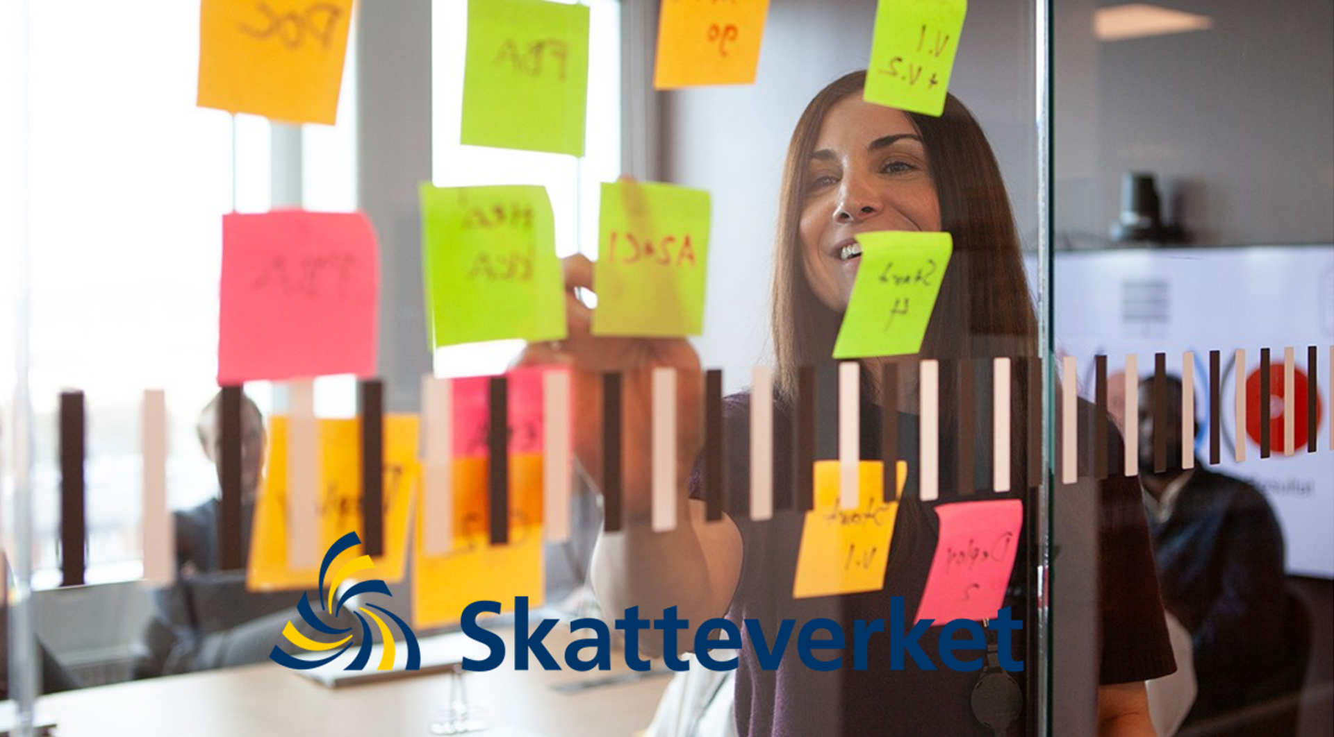 Person sätter upp färgade post-it-lappar på en glasvägg under ett möte eller workshop. Skatteverkets logotyp.